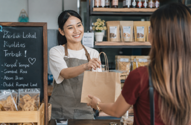 Pemilik UMKM lokal sedang melayani customer di toko kecil dengan produk makanan dan kerajinan lokal Indonesia